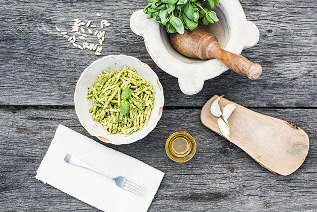 Ein Teller Pasta mit Pesto, dazu Besteck, Pinienkerne und ein Mörser mit Basilikum