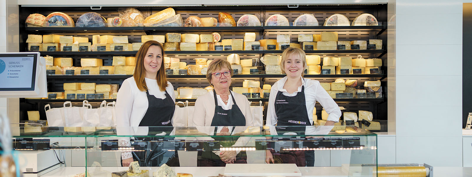 Mercedes Nemeth, Rosa Greiner und Anna Majdic stehen an der Theke im Heiderbeck Feinkost Outlet, im Hintergrund das Käseregal