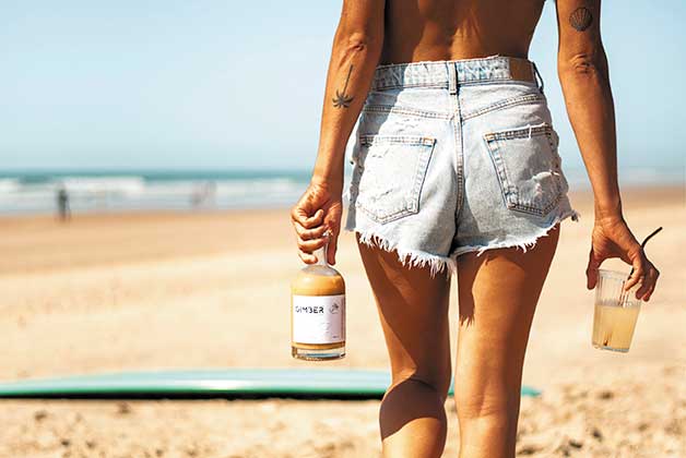 Eine Frau am Strand geht in Richtung mehr und hat eine Flasche Gimber Ingwerdrink in der Hand. In der anderen Hand trägt sie ein Glas