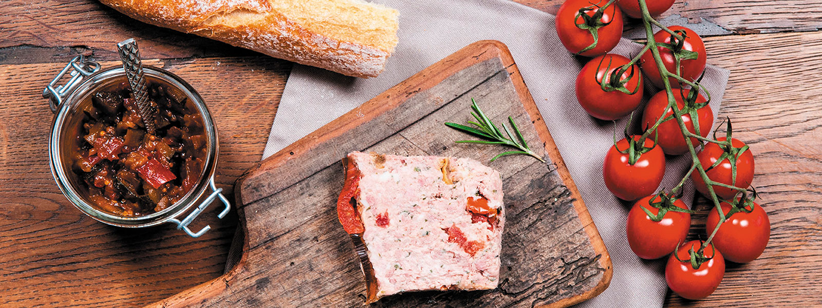 Ein Holztisch, ein angeschnittenes Brot, eine Scheibe Leberpastete auf einem Holzschneidebrett, daneben ein Strauch Tomaten