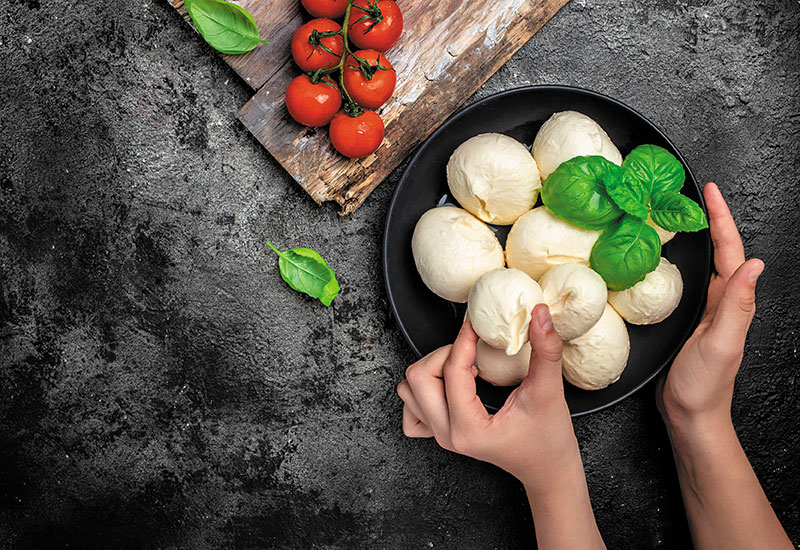 Eine Schale mit Büffelmozzarella Kugeln, Tomaten und frischem Basilikum. Eine Hand zupft die Basililkum Blätter über der Schale