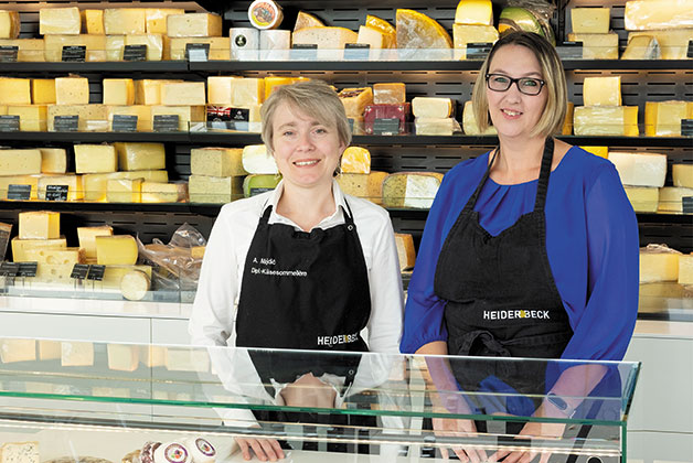 Bild von Anna Maidic und Jennifer Weber die beide als Verkäuferinnen im Käse Outlet arbeiten. Im Hintergrund die Rückwand mit Käse